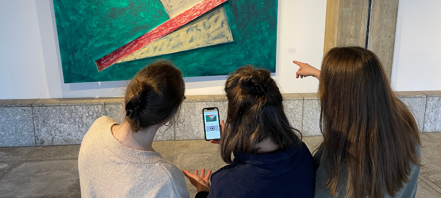 Drei Studierende schauen auf ein Smartphone und schauen sich Informationen zu einem Kunstwerk an.