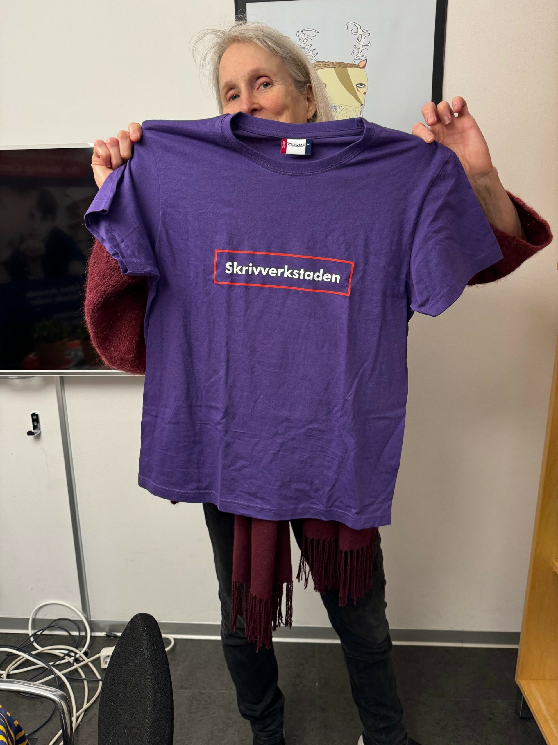 Frau hät ein lilafarbenes T-Shirt mit der Aufschrift "Skrivverkstaden" (dt. Schreibwerkstatt) hoch.