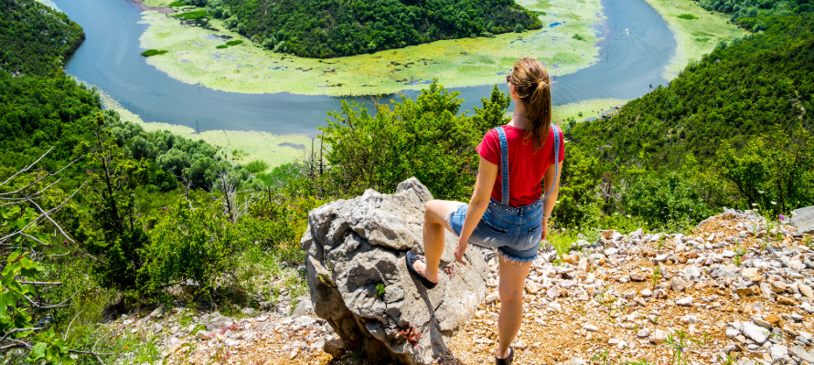 Frau steht mit dem Rücken zum Betrachter auf einem Berg und schaut hinab auf einen von Bäumen umrahmter See.