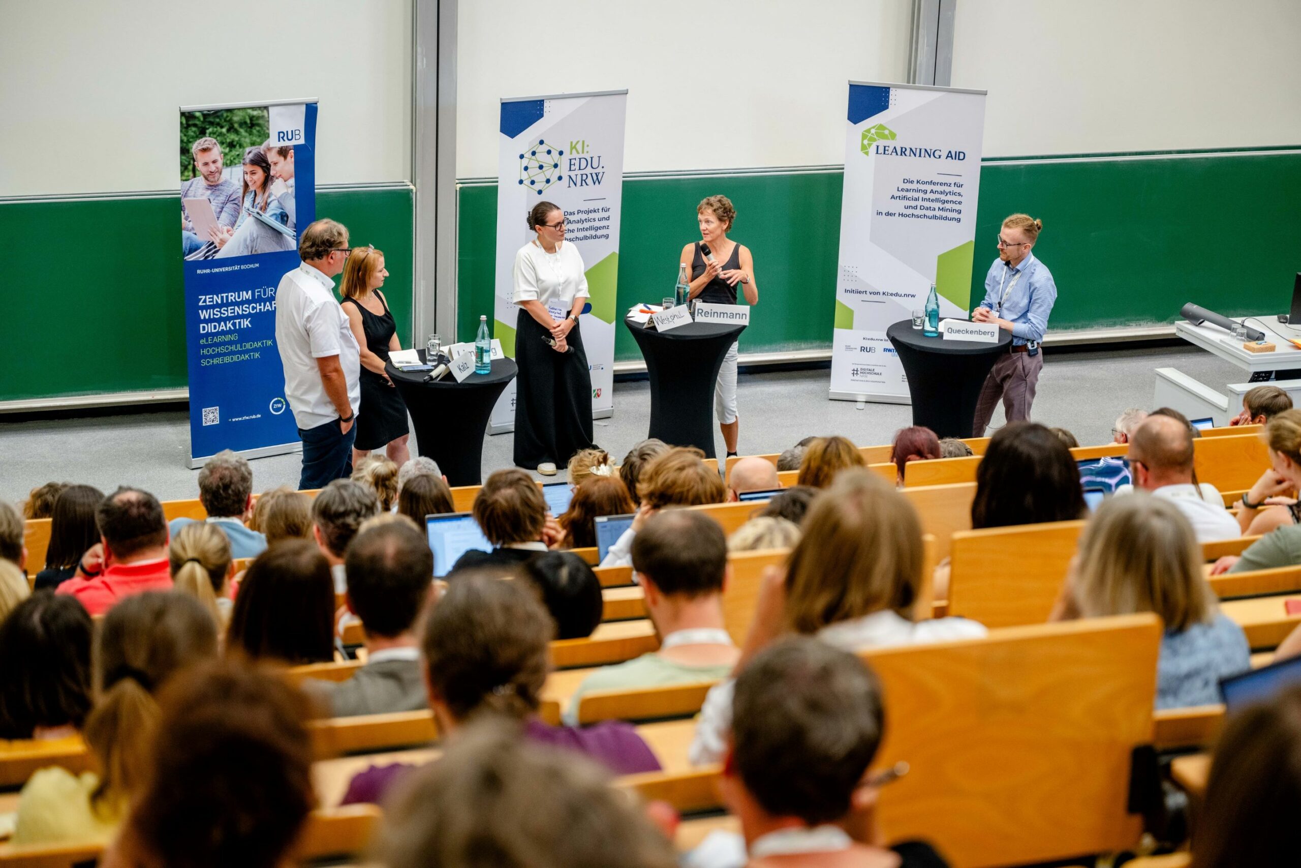 Im Vordergrund sitzen, dem Betrachter mit dem Rücken zugewandt, zahlreiche Personen im Hörsaal. Im Hintergrund befinden sich fünf Personen die eine Diskussion zur Learning AID führen.