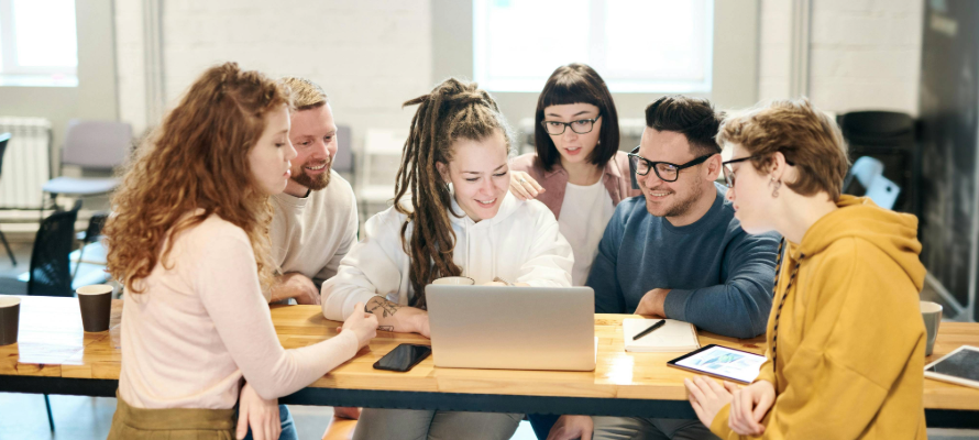 Gruppe von sechse Personen sitzt am Tisch und schaut sich gemeinsam etwas auf einem Laptop an.