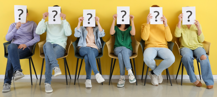 Sechs Personen sitzen auf Stüheln vor einer gelben Wand und halten ein Blatt Papier mit einem großen schwarzen Fragezeichen vor ihren Gesichtern.