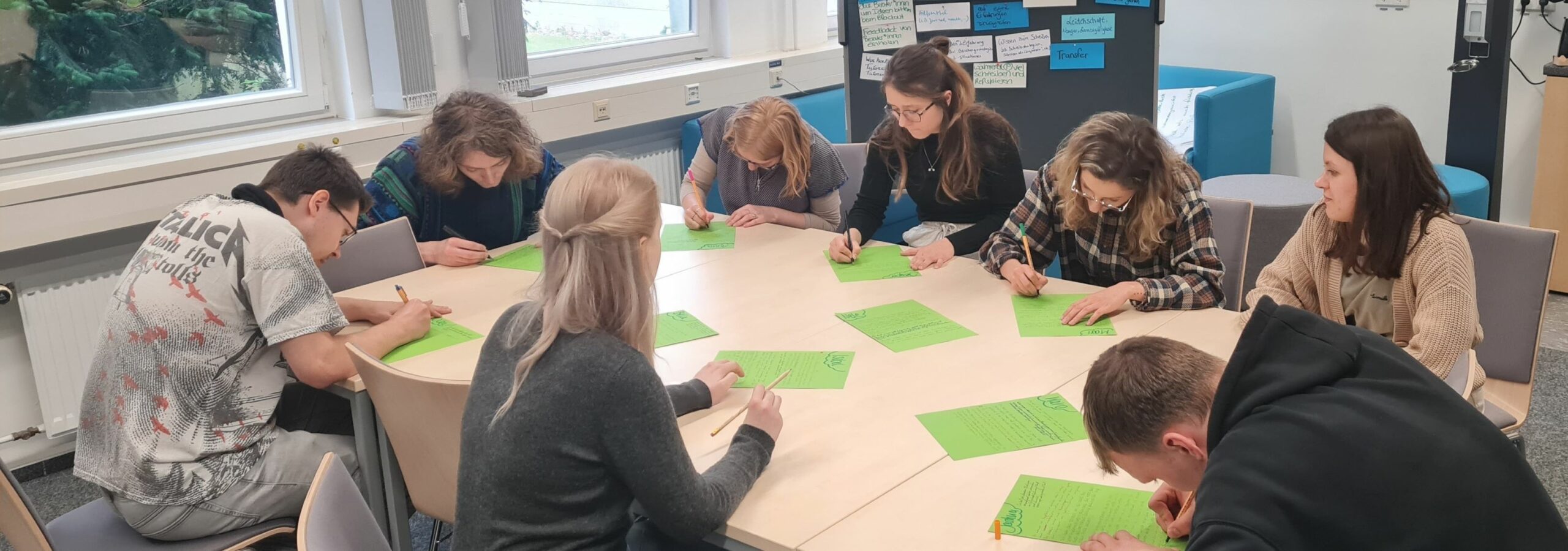 Teilnehmer*innen der Schreibtutor*innen Qualifikation sitzen gemeinsam an einem Tisch und schreiben auf grünen Papierblättern.