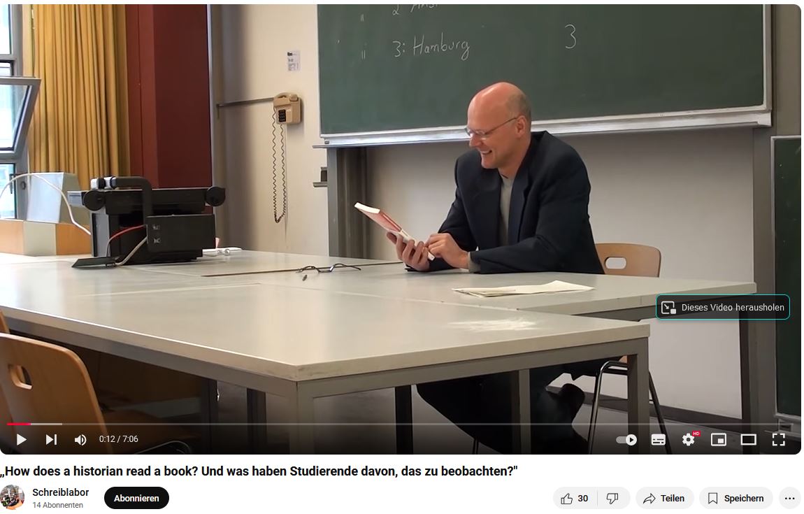 Videoausschnitt: Mann sitzt vor einer Tafel und hält ein Buch in der Hand.