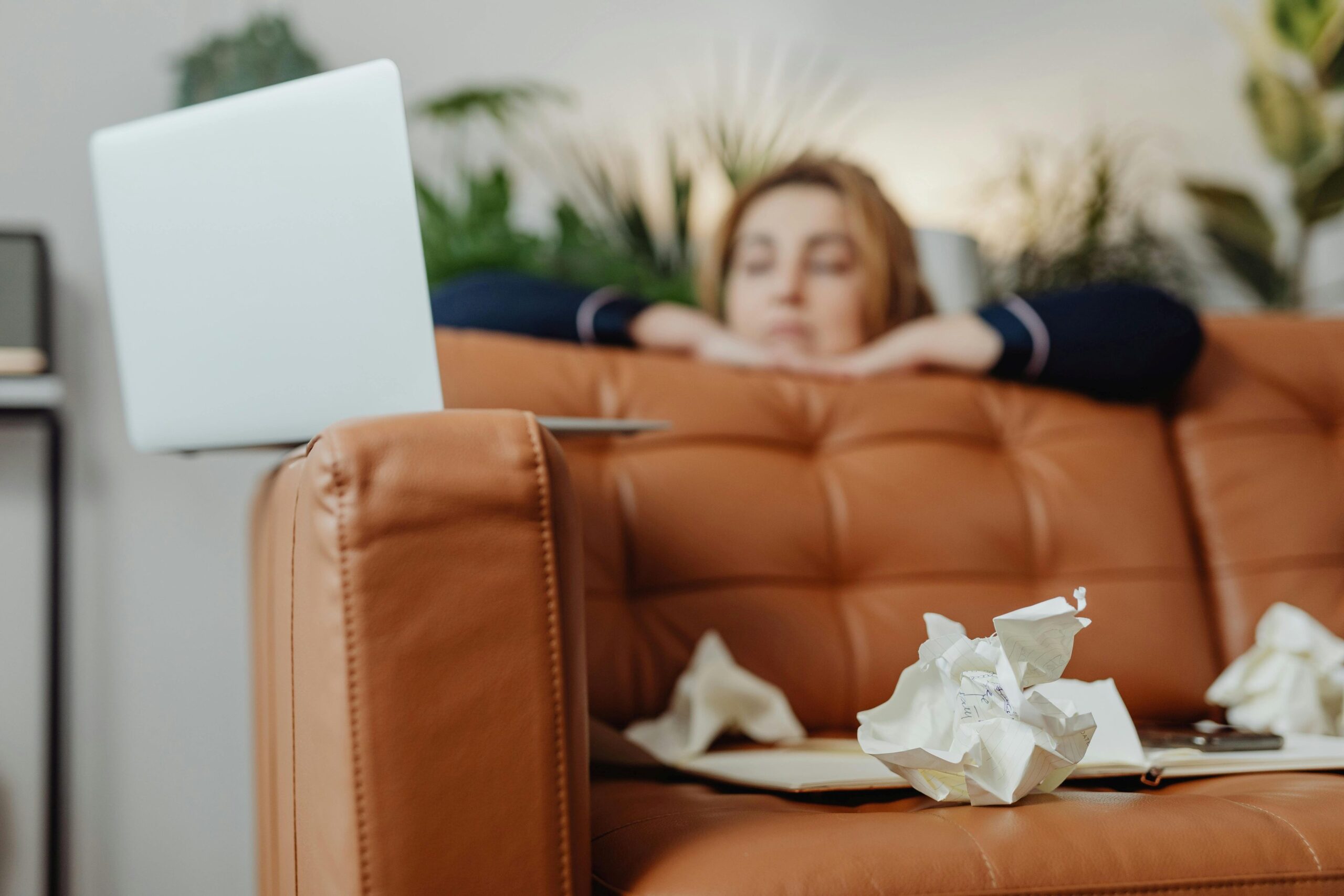 Person lehnt sich auf einem Sofa ab, auf der Sofakante steht ein aufgeklappter Laptop, auf dem Sofasitz liegt zerknülltes Papier.
