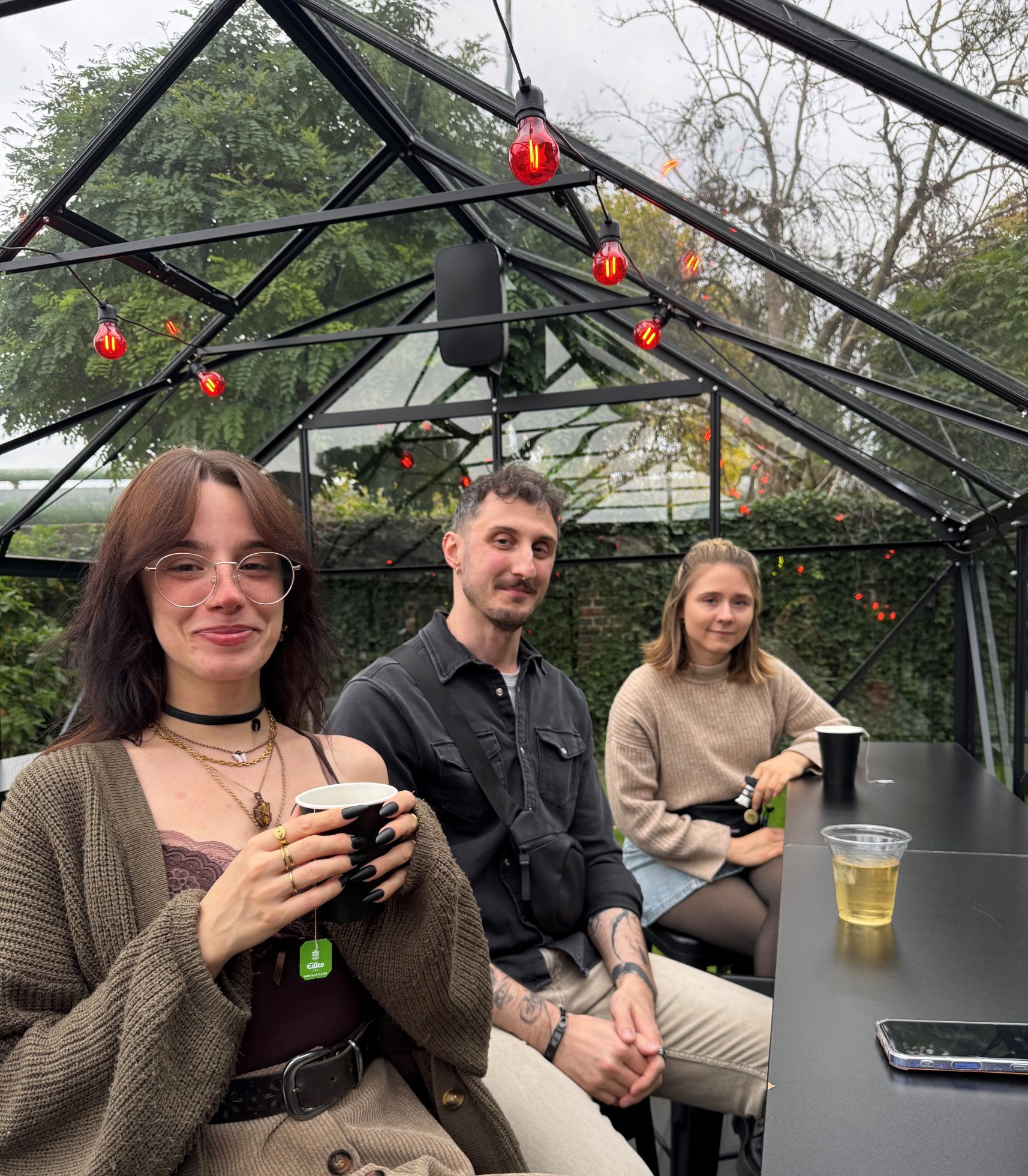 Schreibpeertutor*innen-Konferenz 2025 Bild: 26 C° im Glashäuschen lässt es sich im bewölkten Berlin aushalten (Laura, André und Karo v.l.n.r.).
